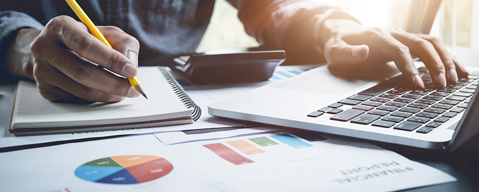 Close-up of a person working on business analysis tasks with a laptop, calculator, notebook and printed charts showing pie and bar graphs.