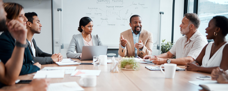 Group of professionals in a conference room engaged in a strategy meeting, with laptops, notebooks, and a whiteboard displaying planning notes.
