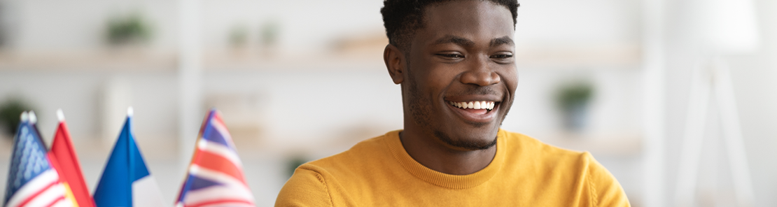  A young man smiles as he learns a new language online. Country flags sit on his desk in a container.